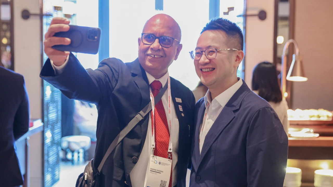 Two men in suits smiling and taking a selfie indoors at a conference or event.