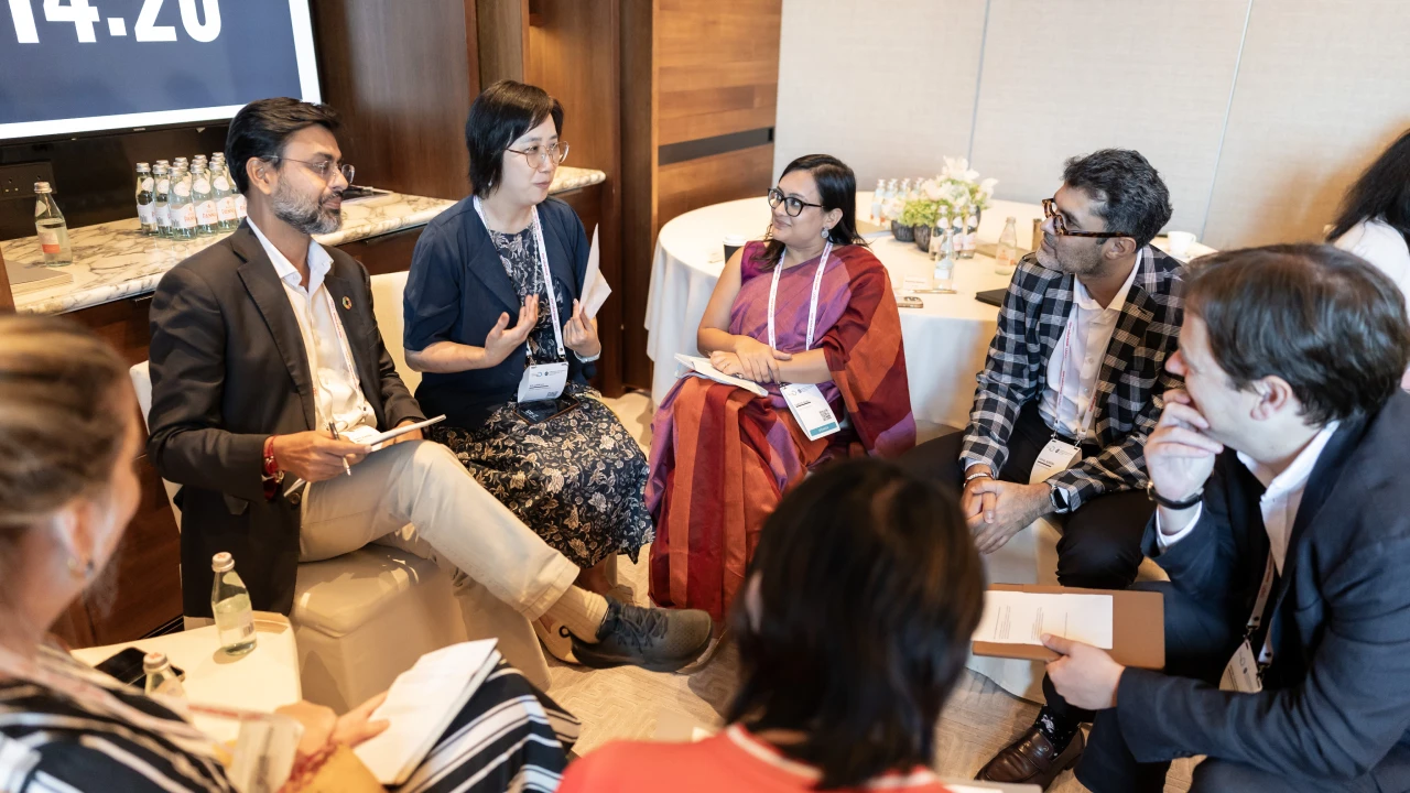 Group of eight diverse people sitting and standing around a low table having a friendly conversation in a green indoor lounge.
